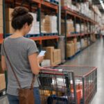 A shopper checking her phone in a warehouse club aisle while pushing a cart